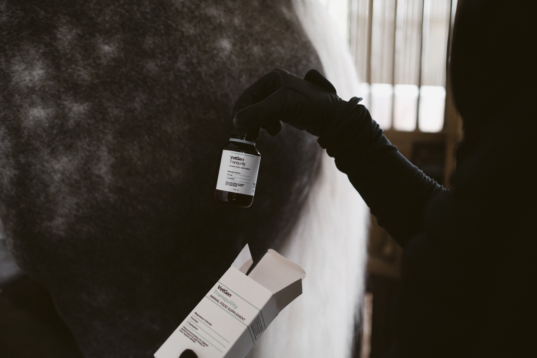 VetGen Tranquility bottle held in a gloved hand beside a grey horse in a stable