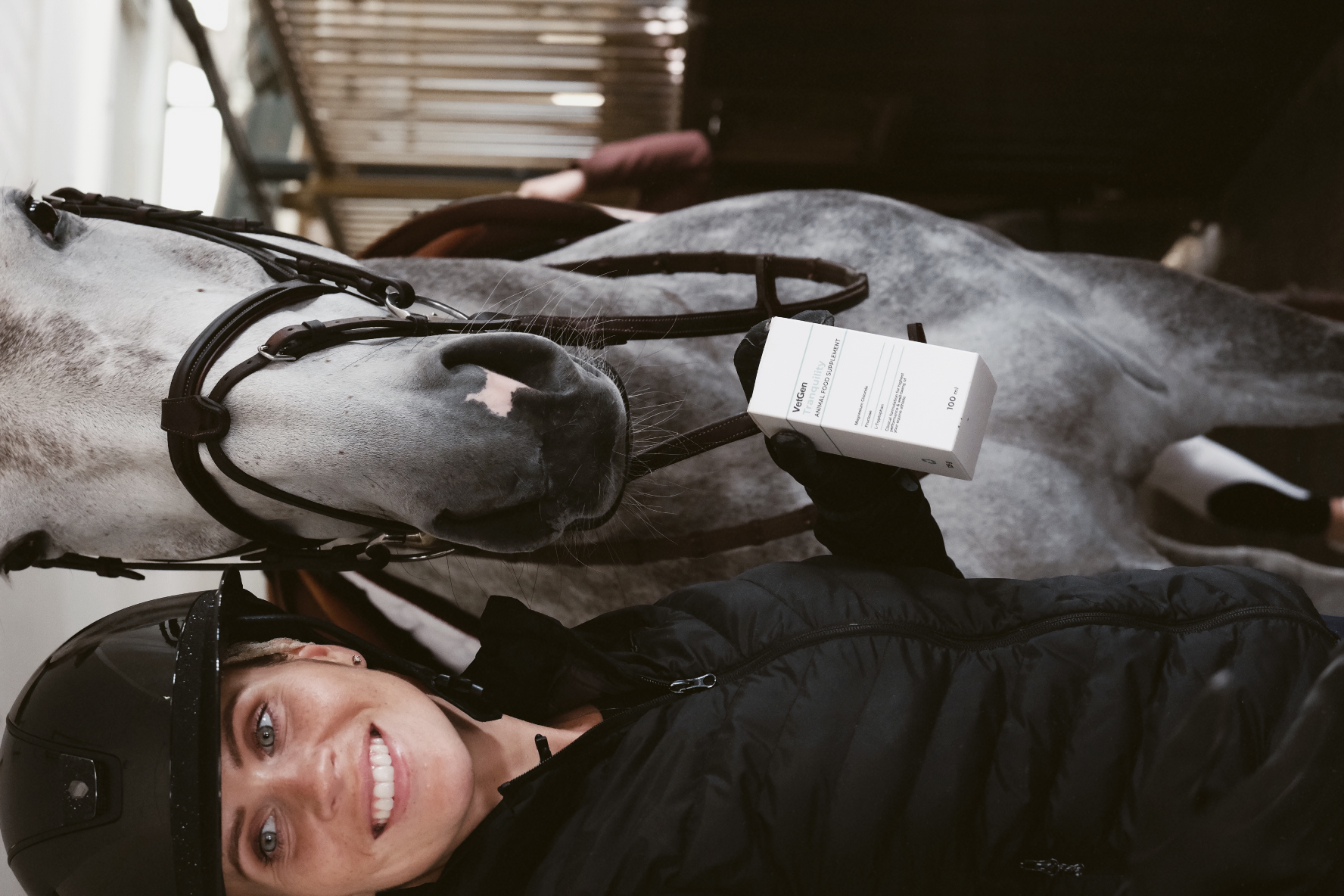 Smiling equestrian in a black helmet and jacket holding a box of VetGen Tranquility beside a dapple-grey horse in a stable